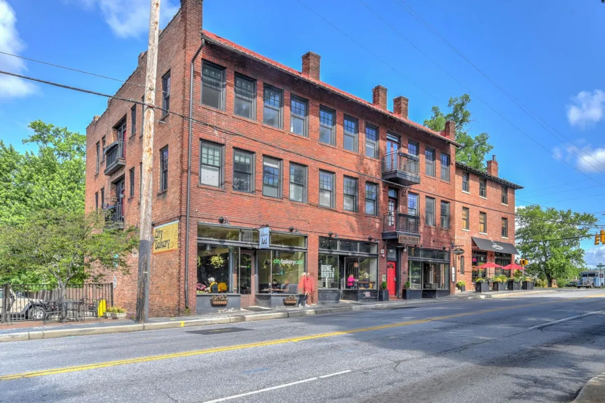 This exterior image features the condo's red brick facade, large windows, and a balcony overlooking lively storefronts at street level. The scene captures a bustling pedestrian-friendly atmosphere with people enjoying nearby boutiques and cafes, illustrating the vibrant community just steps from the residence.