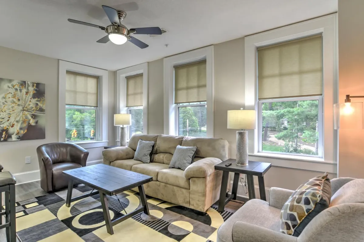 A cozy, sunlit interior living space with a comfortable sofa, coffee table, large windows, and carefully curated artwork. Restored finishes and architectural details are visible, emphasizing the building’s historic charm.