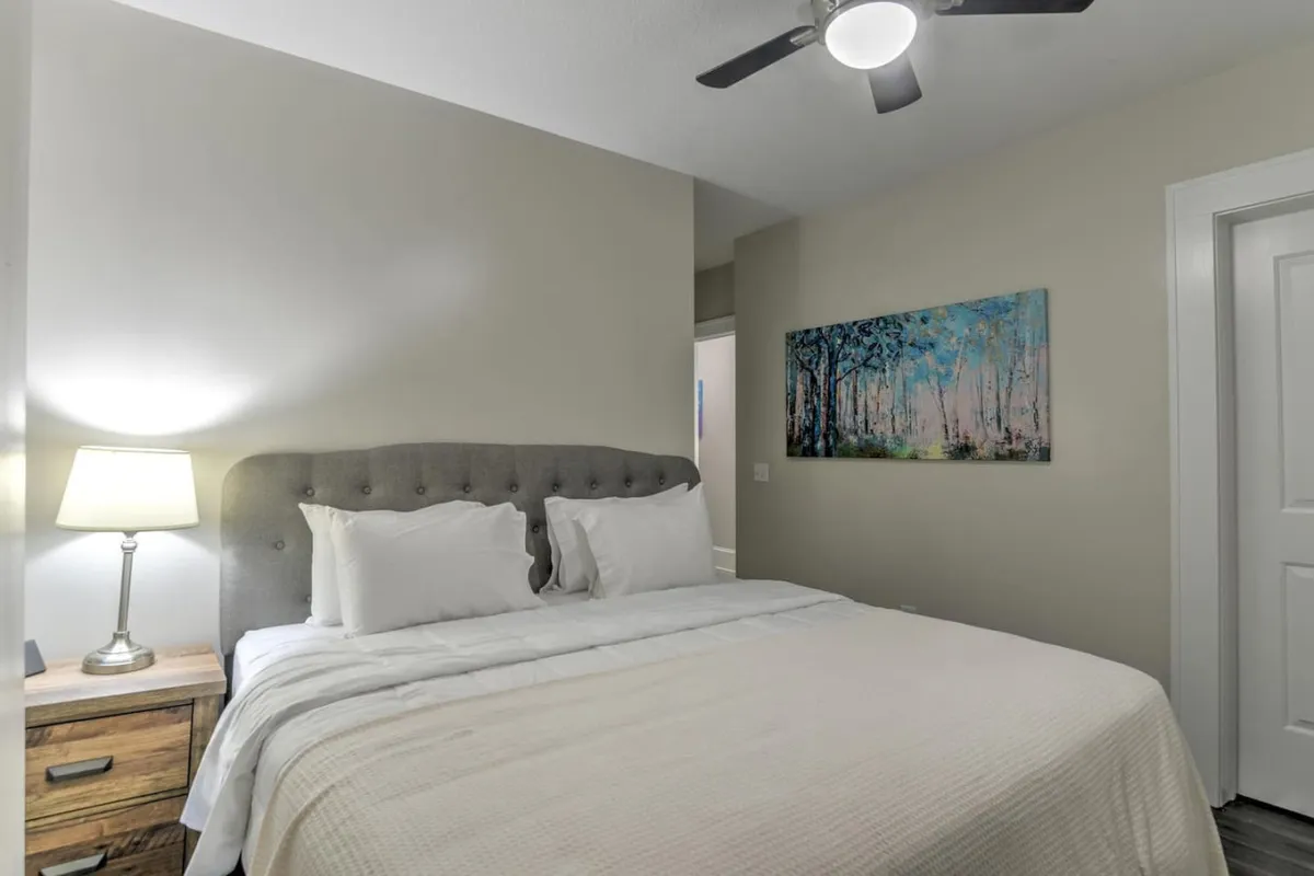 This image displays a tranquil bedroom setting featuring a plush bed adorned with soft bedding, a nightstand with a lamp casting gentle light, and tasteful artwork. The ambiance is serene, evoking a sense of peaceful retreat ideal for restorative rest after a day’s activities.