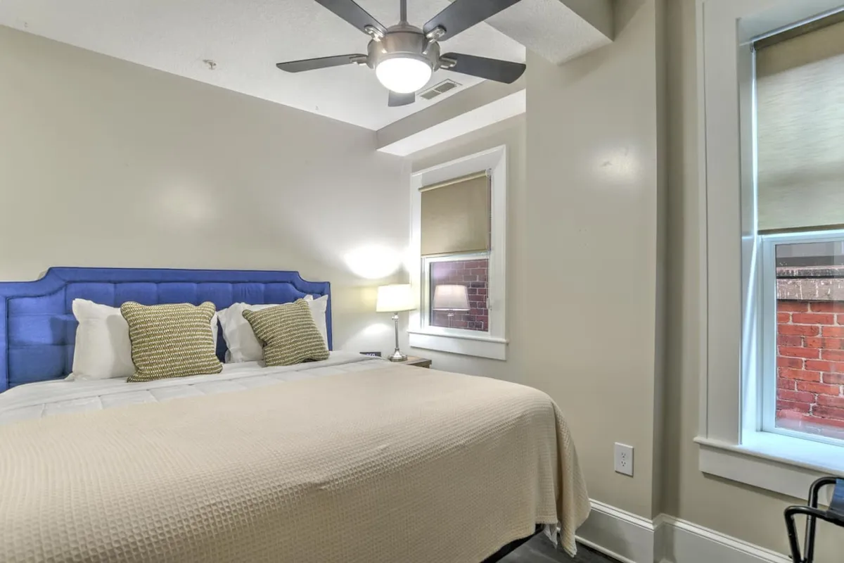 A bright bedroom with a blue upholstered headboard, crisp white linens, decorative pillows, and a bedside lamp. Sunlight pours in through a window, creating a cozy nook perfect for relaxing, reading, or enjoying a quiet morning view.