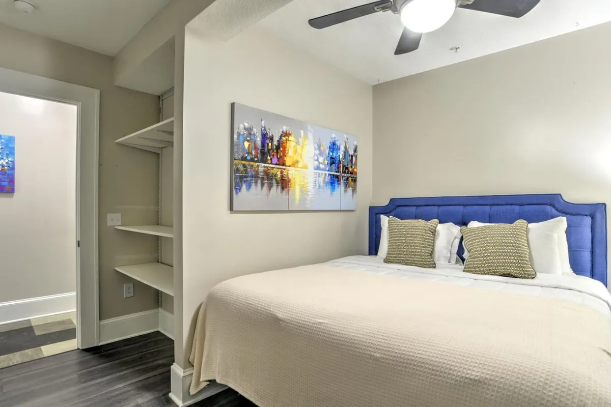 This image showcases a cozy bedroom with a blue upholstered bed, colorful wall art, and a well-organized shelving unit. The room exudes a serene and inviting atmosphere, perfect for relaxation after a day spent exploring the neighborhood or attending business meetings.